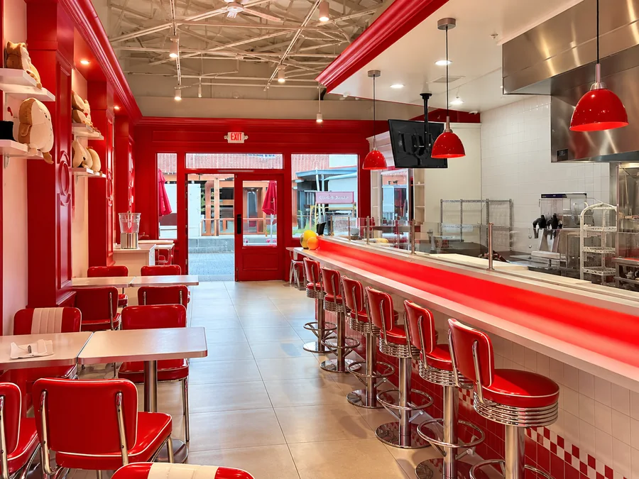 Let’s Toast Restaurant: Finished restaurant dining and bar seating facing the red entry doors; pendants and stainless kitchen pass visible